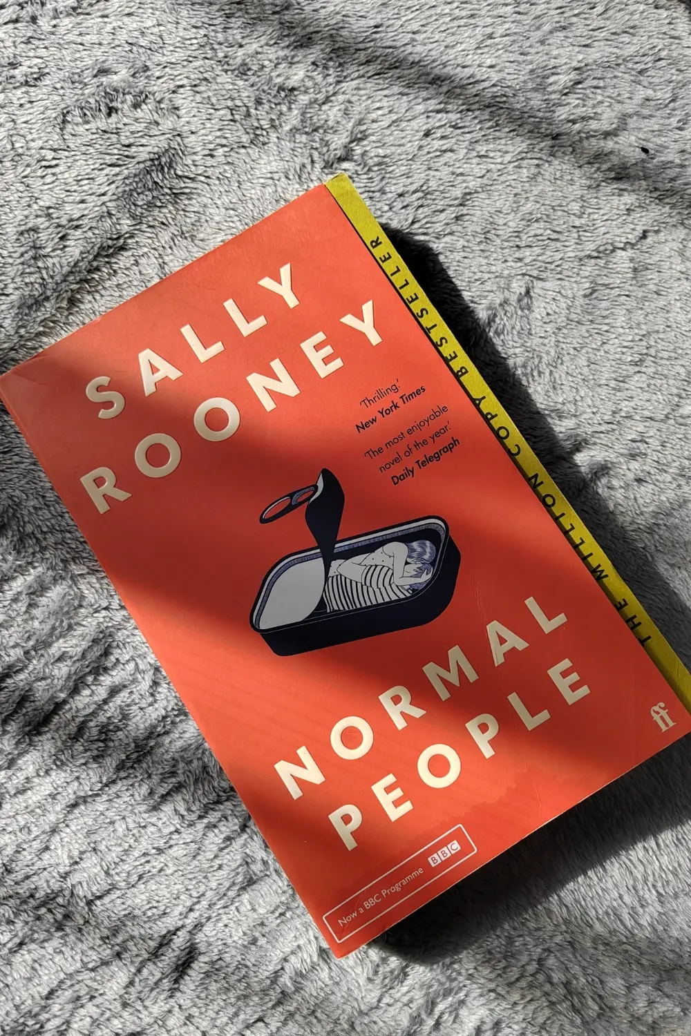 A photo of the paperback copy of Normal People by Sally Rooney against a neutral background (gray blanket). The paperback edition is the red one, with the can of sardines at the front.