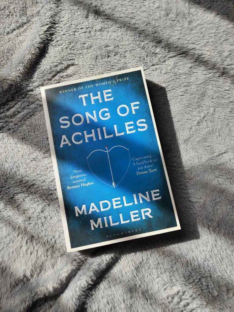 A photo of the paperback edition of The Song of Achilles by Madeline Miller (the blue with an arrow on the front paperback edition), against a gray blanket in the glow of soft sunshine