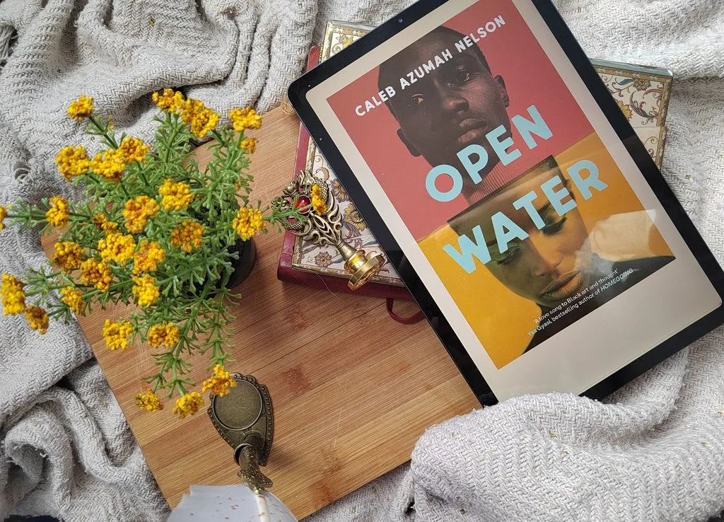 A photo of the e-book edition of Open Water by Caleb Azumah Nelson open on a tablet, set against a flatlay of a golden journal, some yellow wildflowers, a wooden board and a beige blanket.