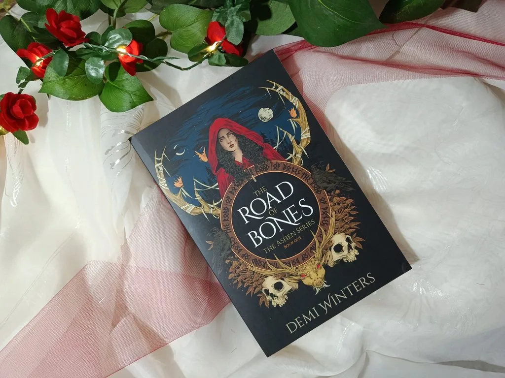 A photo of The Road of Bones by Demi Winters book (paperback edition) against a white background with string lights and roses around it