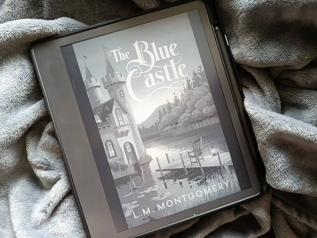 A photo of The Blue Castle by L. M. Montgomery open on a Kindle Scribe so it's showing the cover, against a soft gray blanket