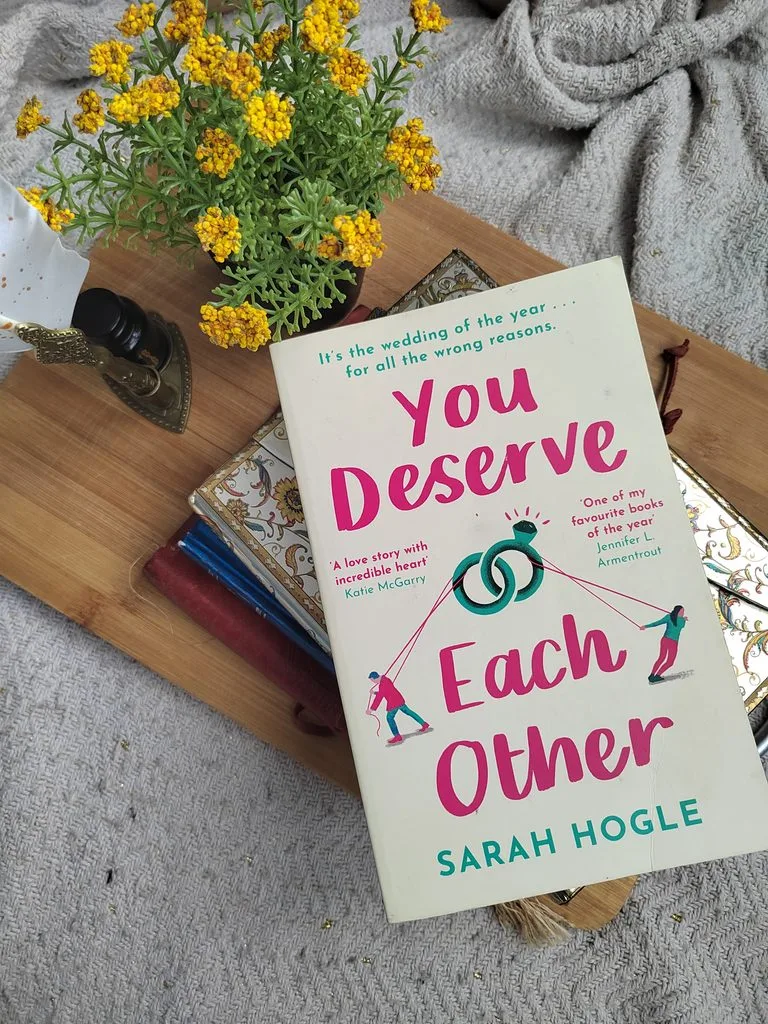 A photo of the paperback edition (UK) of You Deserve Each Other by Sarah Hogle, against a background of some golden, red and blue journals, a wooden board and some yellow wildflowers