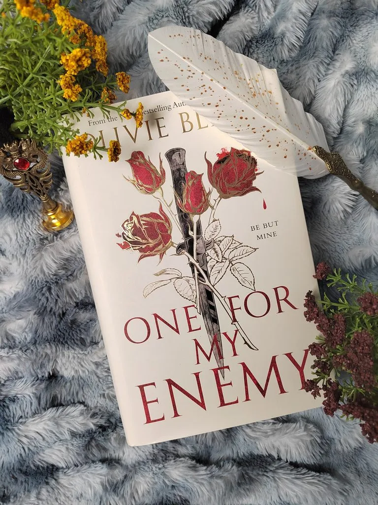 A photo of the illustrated hardback edition of One of My Enemy by Olivie Blake with some wildflowers in red and yellow against a bluish gray blanket