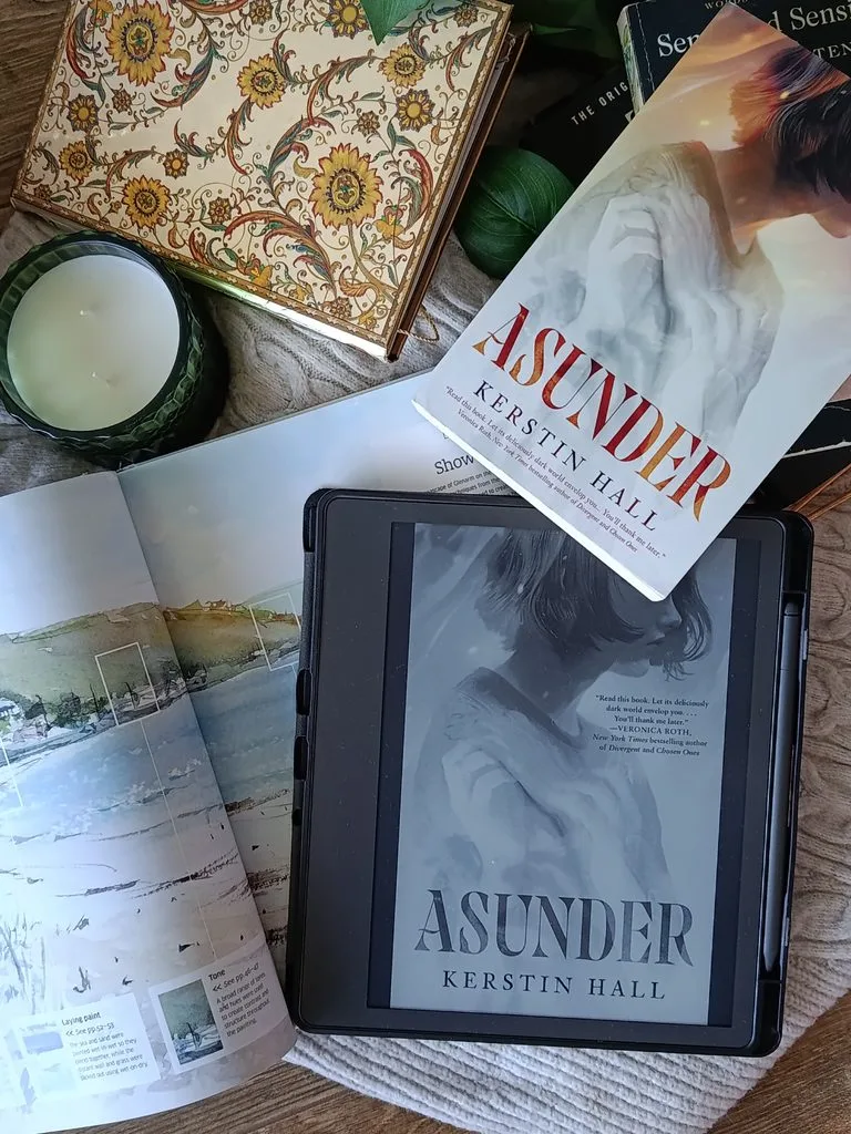A photo showing the paperback edition of Asunder by Kerstin Hall and the e-book version of Asunder by Kerstin Hall open on Kindle Scribe so the cover can be seen